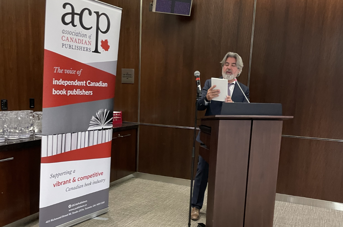 Minister of Canadian Heritage Pablo Rodriguez standing 
                     at a podium while holding a blank book
                     